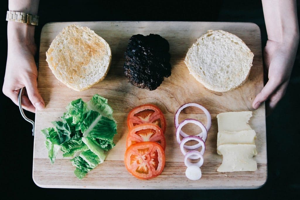 hamburger with hamburger buns on a cutting board with red tomato, green lettuce, red onion, cheese