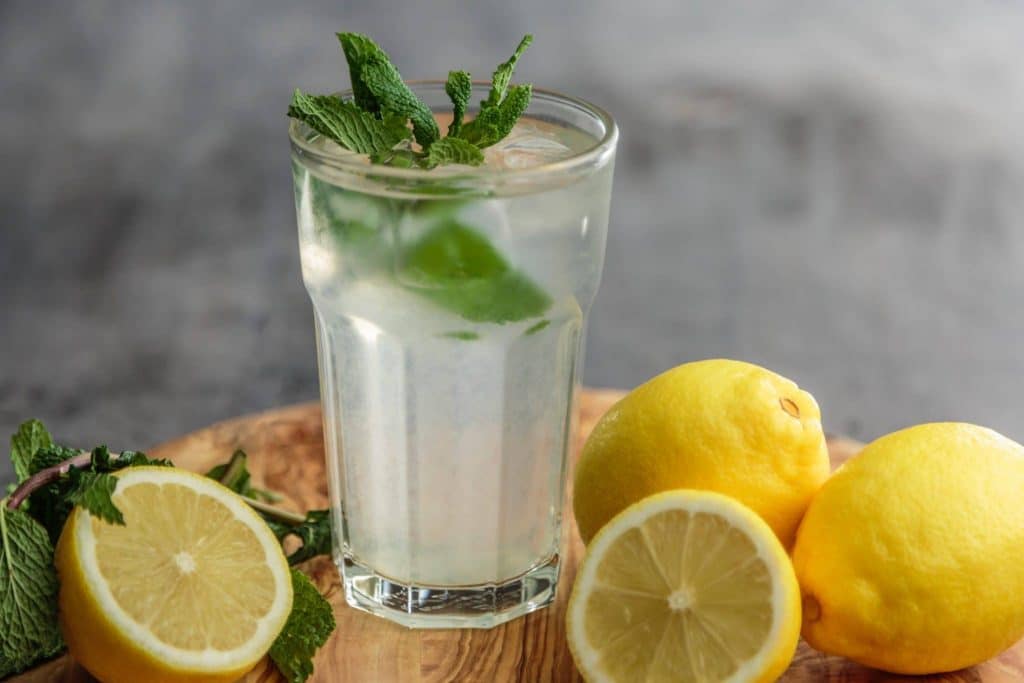 lemon water with mint leaves in clear glass with sliced yellow lemons nearby