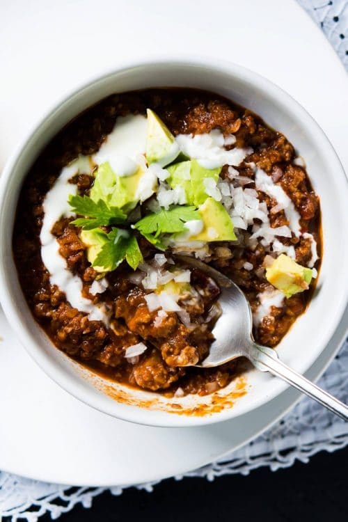 overhead shot chili in a white bowl with avocado chunks, sour cream, onions, and cilantro with spoon