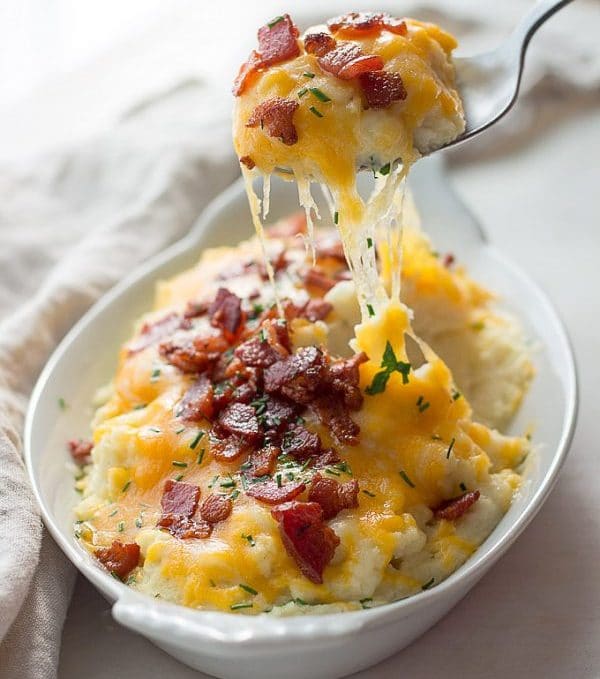 mashed cauliflower in white serving dish topped with bacon, chives, and shredded cheddar cheese