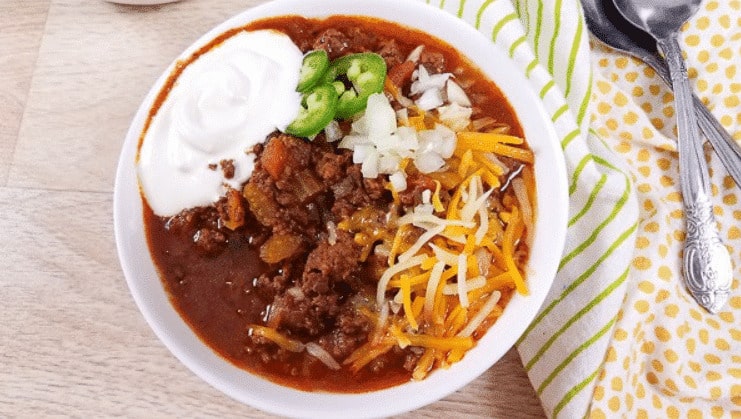 chili topped with shredded cheddar cheese, onions, jalapenos, and sour cream in a white bowl with spoons nearby