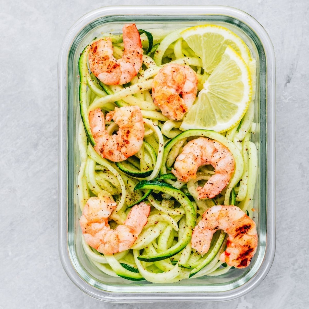 Overhead view of shrimp zoodles meal prep with lemon and lime wedges in a glass container.