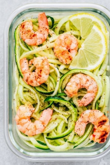 Overhead view of shrimp zoodles meal prep with lemon and lime wedges in a glass container.