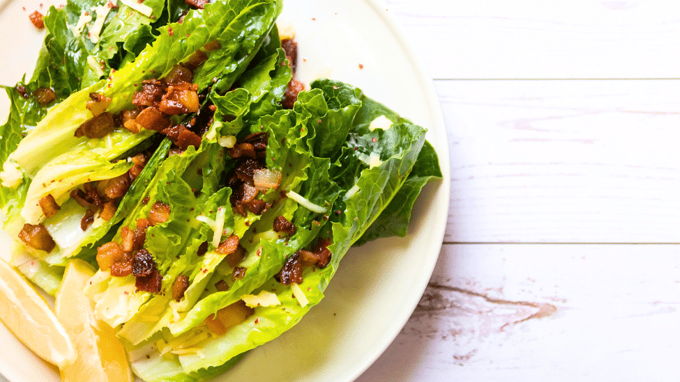 romaine lettuce leaves with bacon bits on top, lemon on the side, on a white plate on a white wooden table