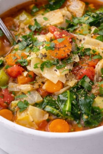 Close-up of keto cabbage soup in a white bowl with visible cabbage, carrots, tomatoes, and fresh parsley