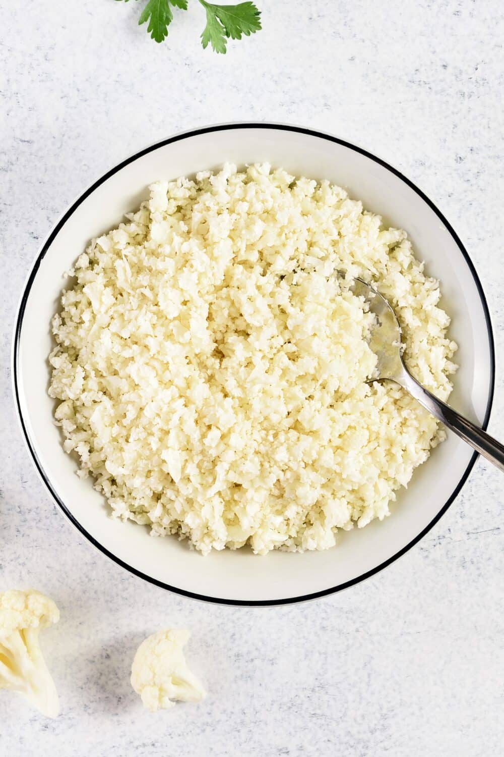 Overhead shot of finely grated cauliflower rice presented in a white bowl with a sleek black outline around the rim, contrasted against fresh sprigs of parsley nearby, highlighting a healthy, keto-friendly alternative to traditional rice