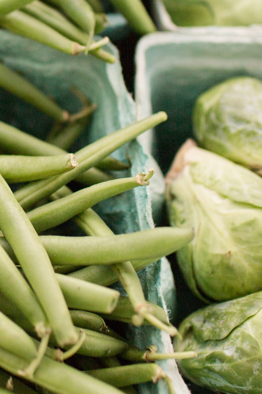 Fresh, green Brussels sprouts and slender green beans showcased side by side in recyclable green paper containers at a local market, highlighting the organic and healthy produce available for purchase.