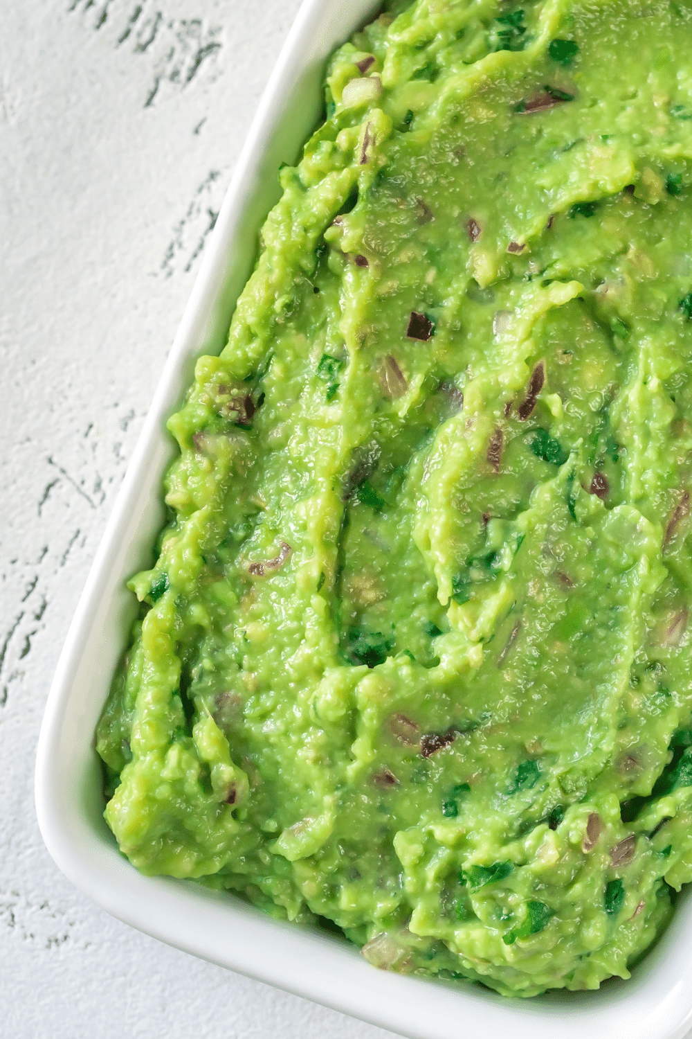 An appetizing serving of guacamole is elegantly presented in a square white dish, resting atop a smooth white wooden table. The vibrant green guacamole is beautifully textured, showcasing a luscious blend of creamy avocado, diced tomatoes, finely chopped onions, and aromatic cilantro. The guacamole's velvety surface is artfully adorned with a scattering of ruby-red pomegranate arils, adding a burst of color and a touch of fruity sweetness. Surrounding the guacamole dish are a few crisp tortilla chips, strategically arranged to invite sampling. The white dish's clean lines and minimalist design provide a perfect canvas for the guacamole's vibrant hues to shine. The dish sits harmoniously on the pristine white wooden table, emphasizing a sense of freshness and natural appeal. Sunlight gently bathes the ensemble, casting soft shadows that further accentuate the dish's inviting presentation. The sight of this guacamole creation promises a delightful taste experience, inviting all to indulge in the rich flavors and satisfying textures of this classic and beloved dip.