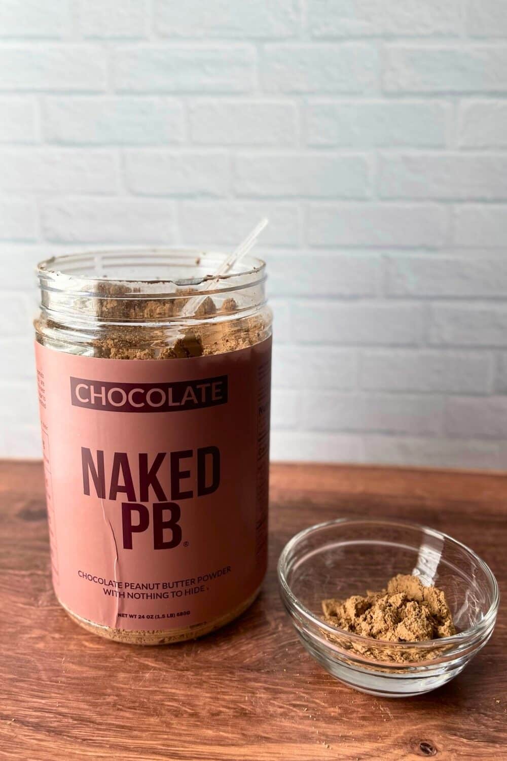 Revealing its contents, the Naked Nutrition Chocolate Powdered Peanut Butter container stands open, its lid resting beside it. A sturdy scoop rests atop the rich cocoa-infused peanut powder, inviting culinary creativity. Nearby, a petite glass container holds an additional scoop, poised for action. The scene unfolds against a backdrop of a clean white surface and a rustic wooden table.