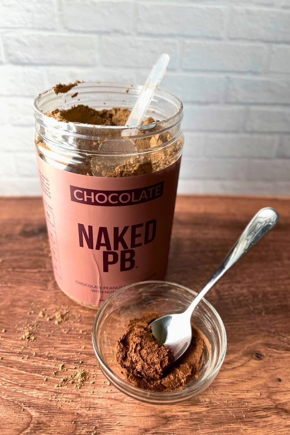 Unveiling the transformation, the Naked Nutrition Chocolate Powdered Peanut Butter jar stands open, its scoop nestled atop the velvety brown powder. Adjacent, a petite glass container holds a scoop transformed into a luscious peanut butter paste, having been reconstituted with a touch of water. This captivating tableau unfolds against a backdrop of a pure white surface and a natural wooden table.