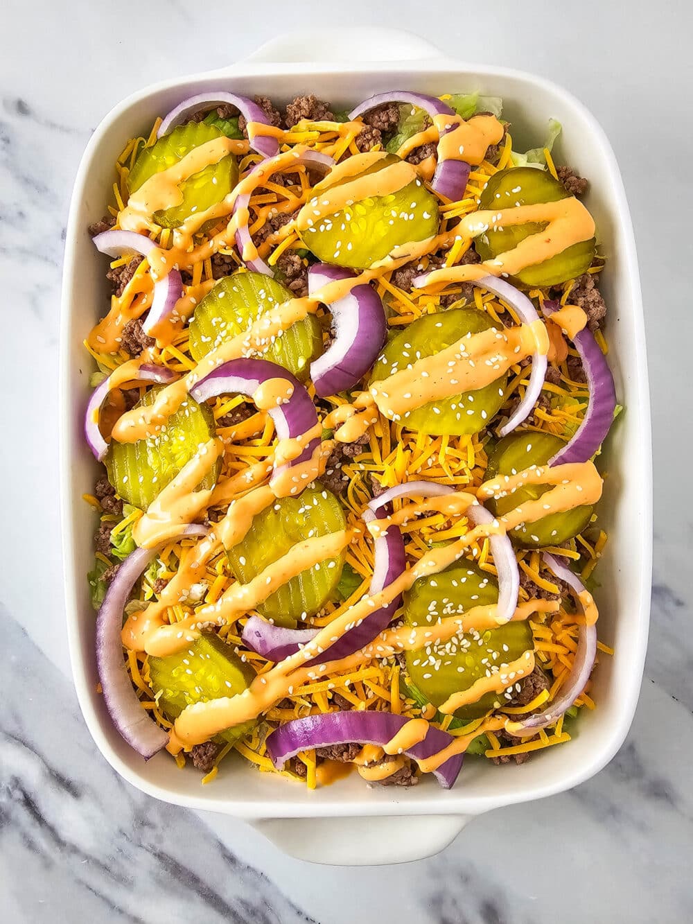 chopped lettuce and ground beef and shredded cheese and pickles and onions and thousand island dressing and sesame seeds in a casserole dish