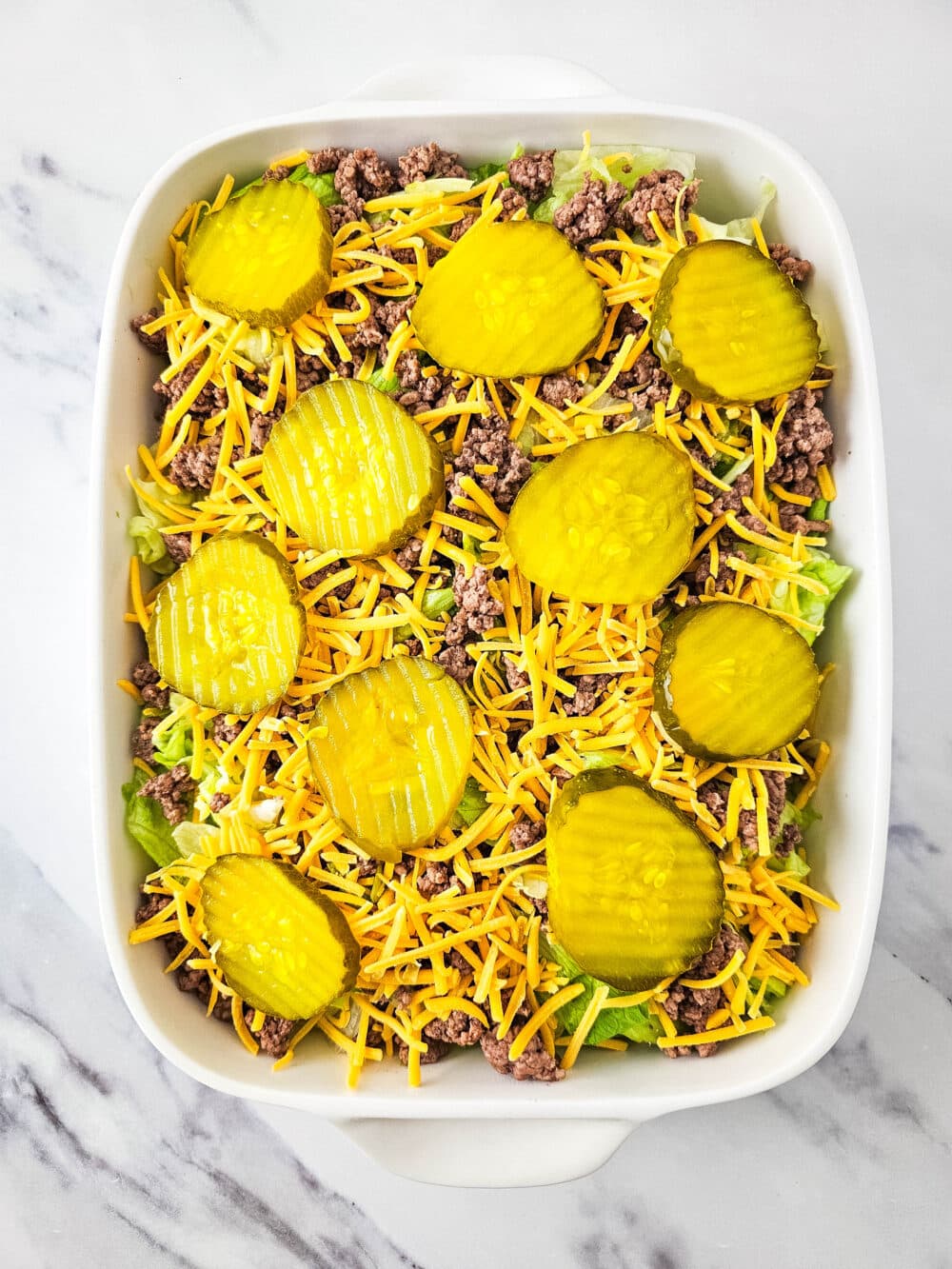 chopped lettuce and ground beef and shredded cheese and pickles in a casserole dish
