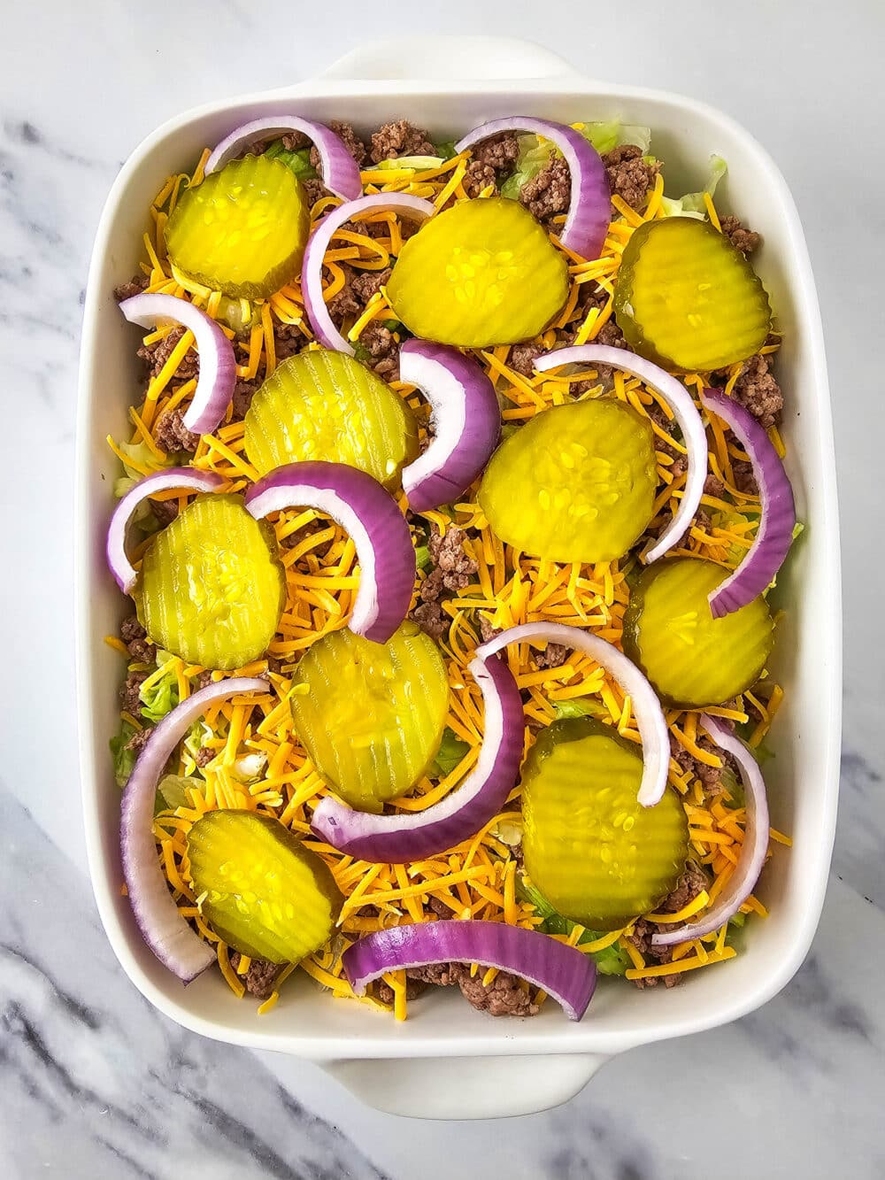 chopped lettuce and ground beef and shredded cheese and pickles and onions in a casserole dish