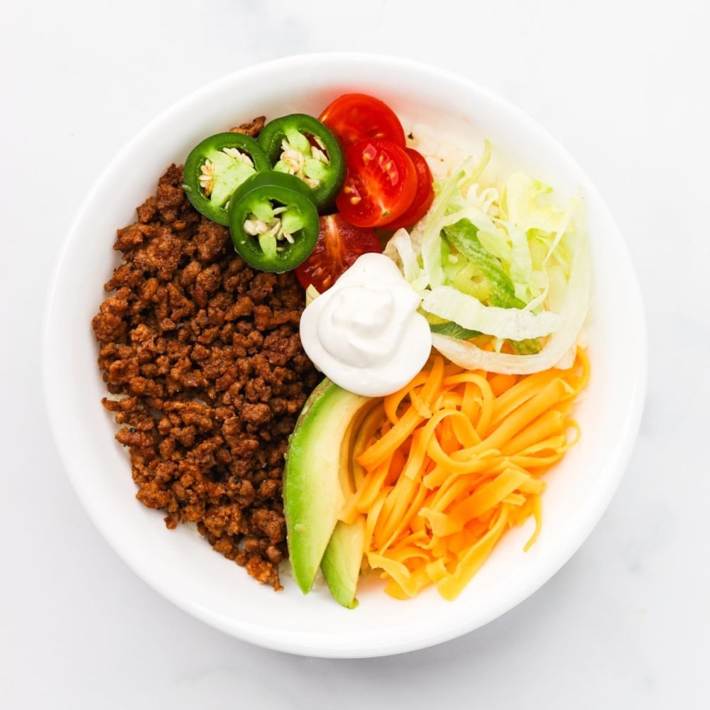 overhead shot of keto taco bowls topped with avocado, shredded lettuce, shredded yellow cheese, sliced jalapenos and cherry tomatoes