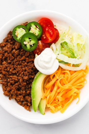 overhead shot of keto taco bowls topped with avocado, shredded lettuce, shredded yellow cheese, sliced jalapenos and cherry tomatoes