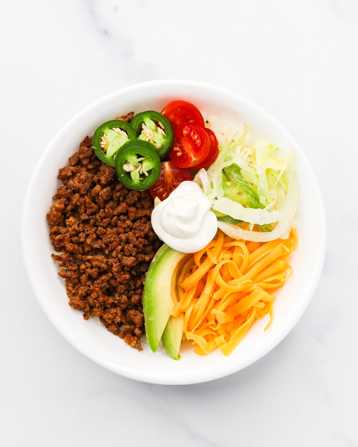 High protein keto taco bowl with ground beef, shredded cheddar, avocado, cherry tomatoes, and jalape&ntilde;os in a white bowl