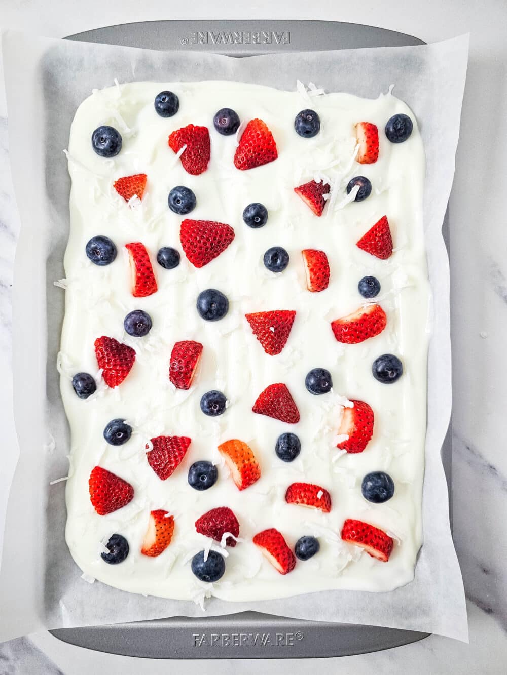 strawberries and blueberries and unsweetened coconut flakes on greek yogurt on a baking pan