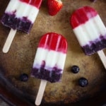 3 red, white, and blue popsicles on a pan with strawberry and blueberries nearby