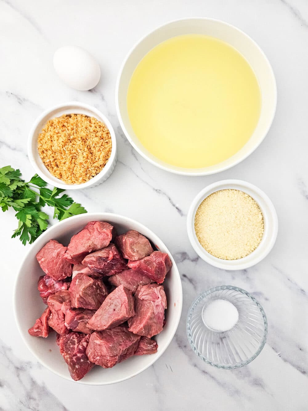 Ingredients laid out on a marble surface including raw steak chunks, egg, grated parmesan, crushed pork rinds, oil, parsley, and salt.