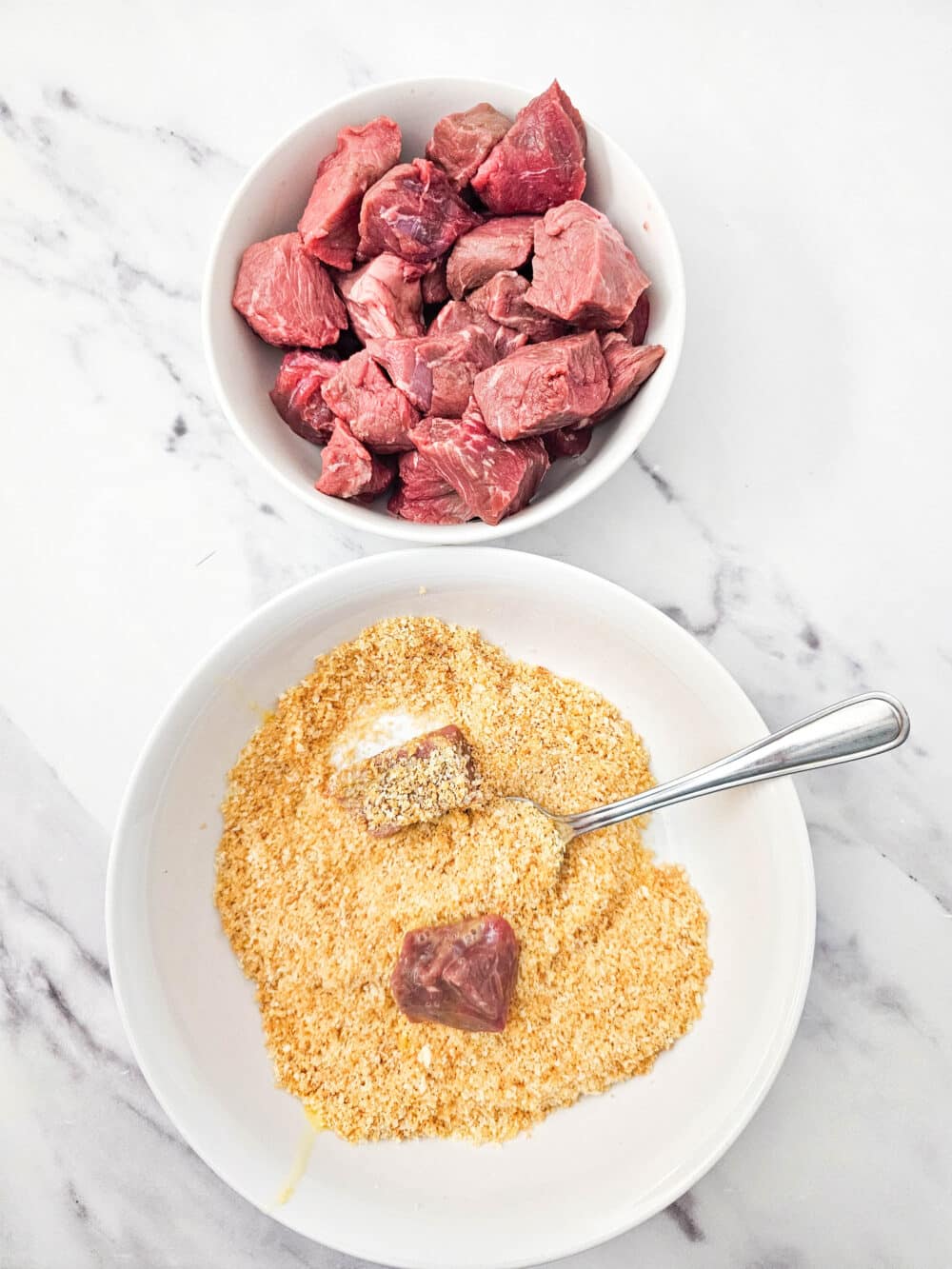 Raw steak chunks being coated in a pork rind and parmesan breading mixture.