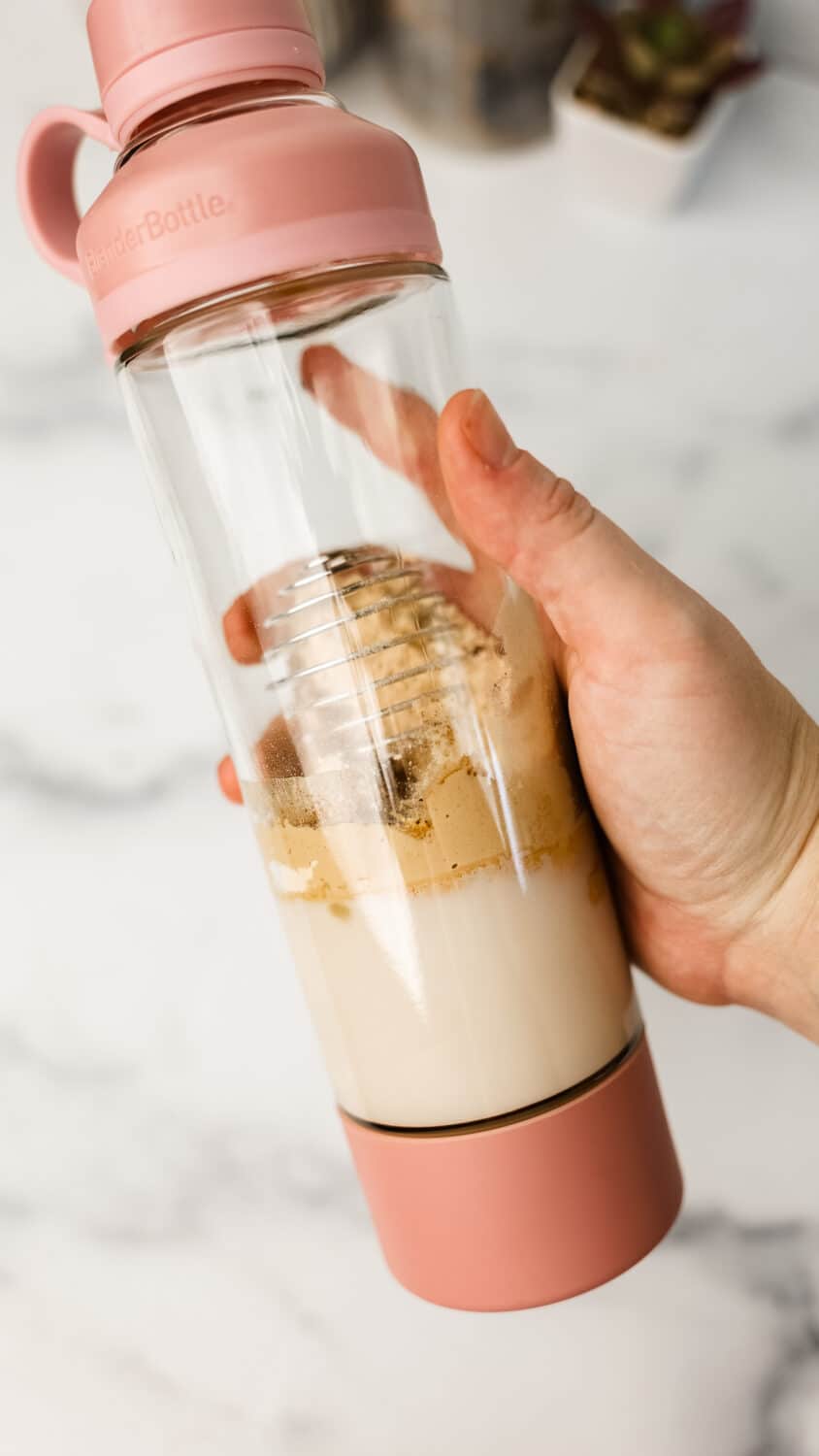 Hand holding a pink blender bottle filled with almond milk, protein powder, and spices before shaking.