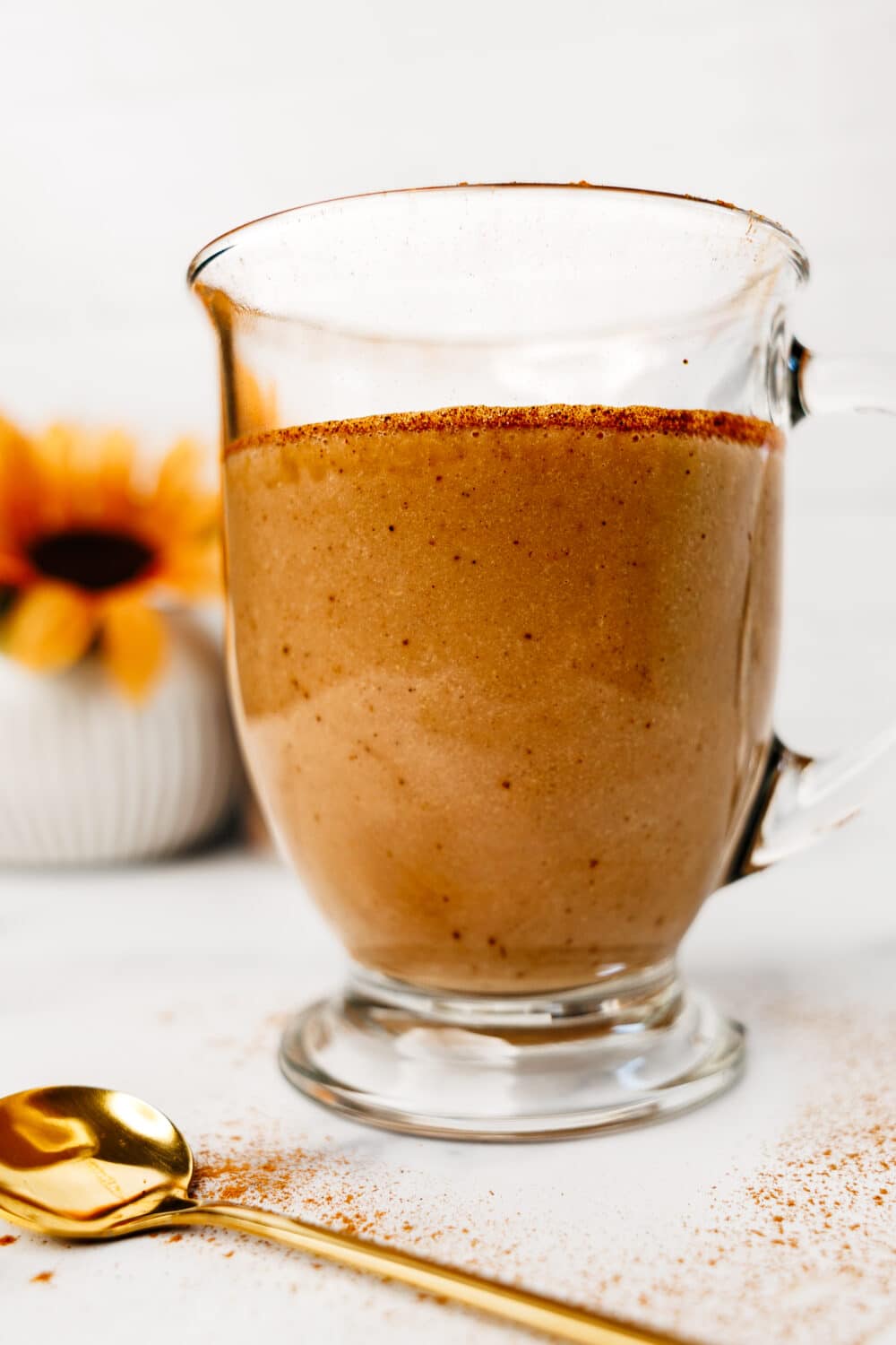 Side view of a glass mug filled with protein chai latte, sprinkled with cinnamon, with a gold spoon and sunflower in the background.