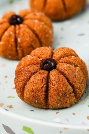 Pumpkin-shaped energy bites with ridged sides and a chocolate chip stem, arranged on a speckled plate.