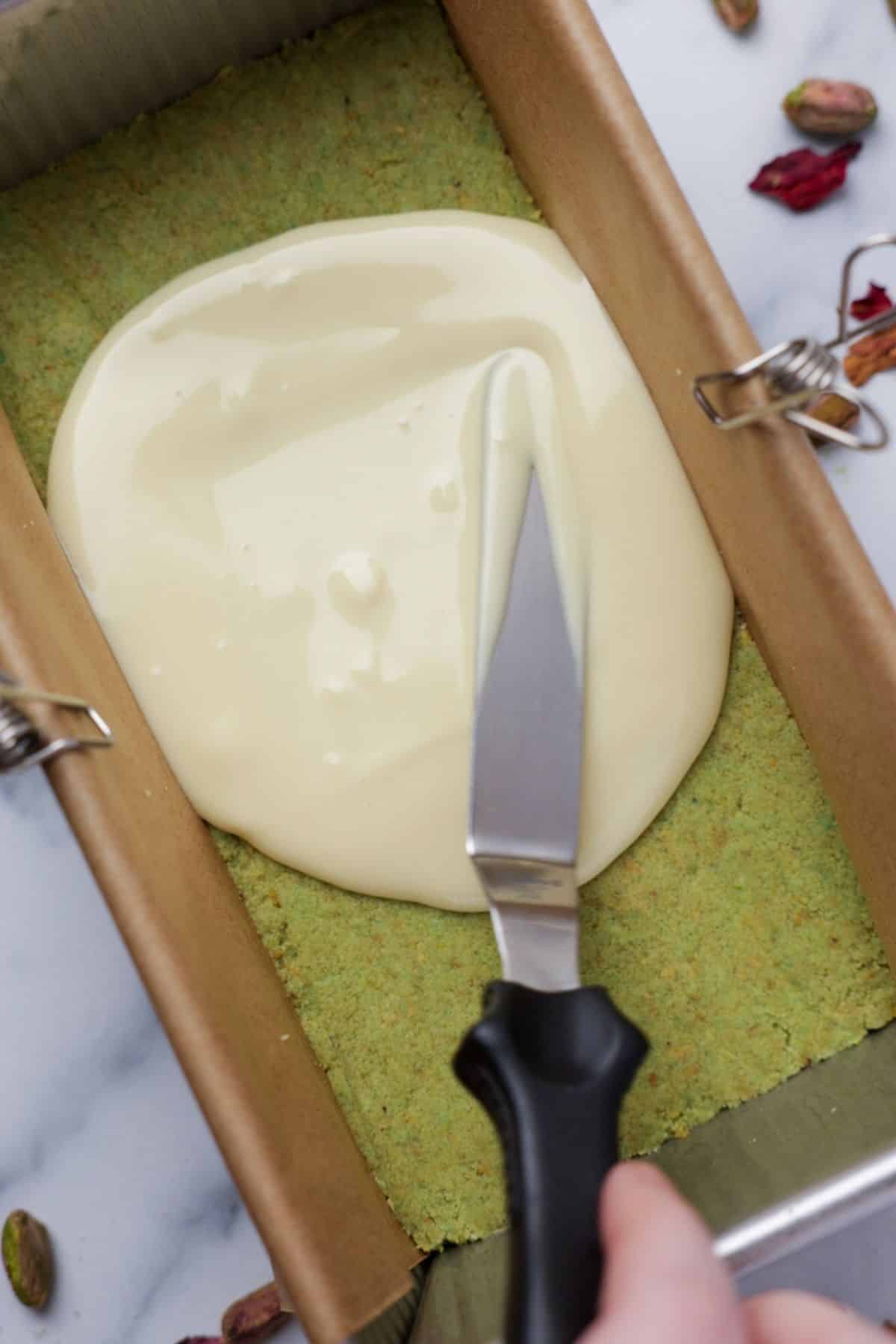 Melted white chocolate being spread over pistachio base in loaf pan using offset spatula