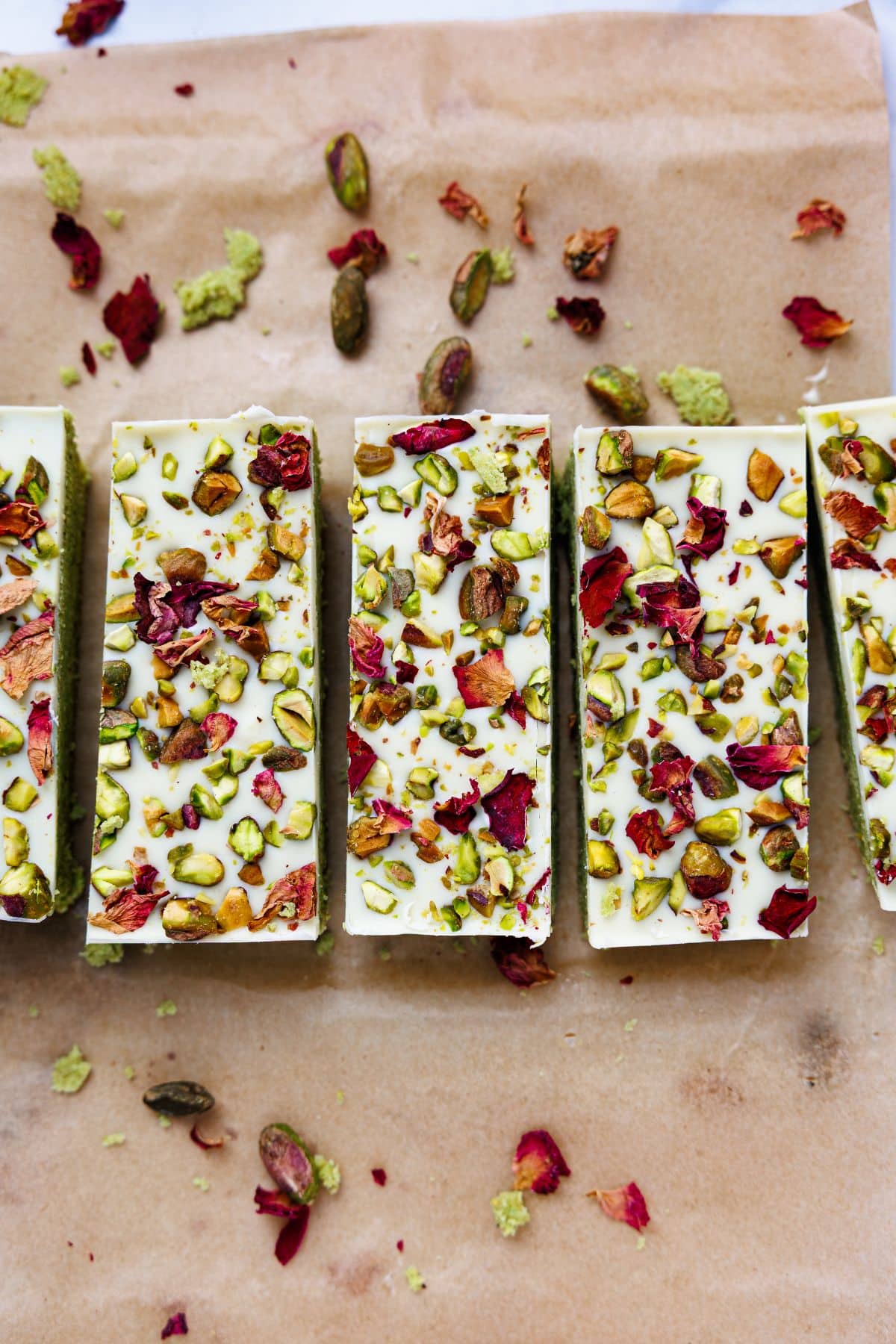 Four pistachio protein bars arranged in row on parchment paper, showing white chocolate tops decorated with pistachios and rose petals