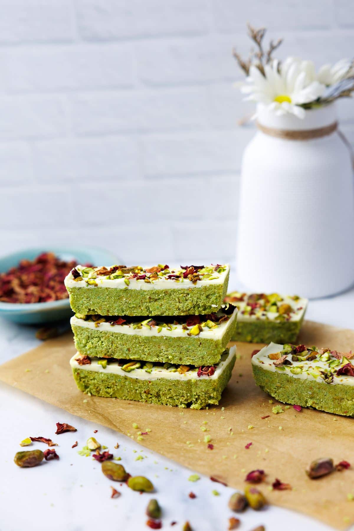 Stack of three pistachio protein bars on parchment paper with white ceramic vase and daisies in soft-focused background