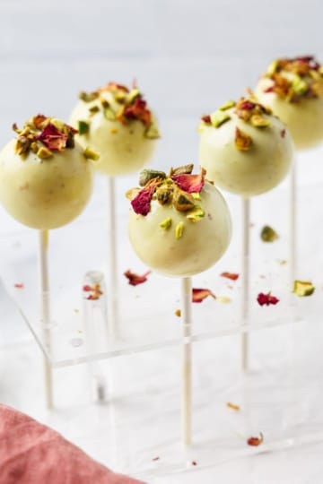 Pistachio cake pops coated in white chocolate topped with chopped pistachios and dried rose petals displayed in acrylic cake pop stand