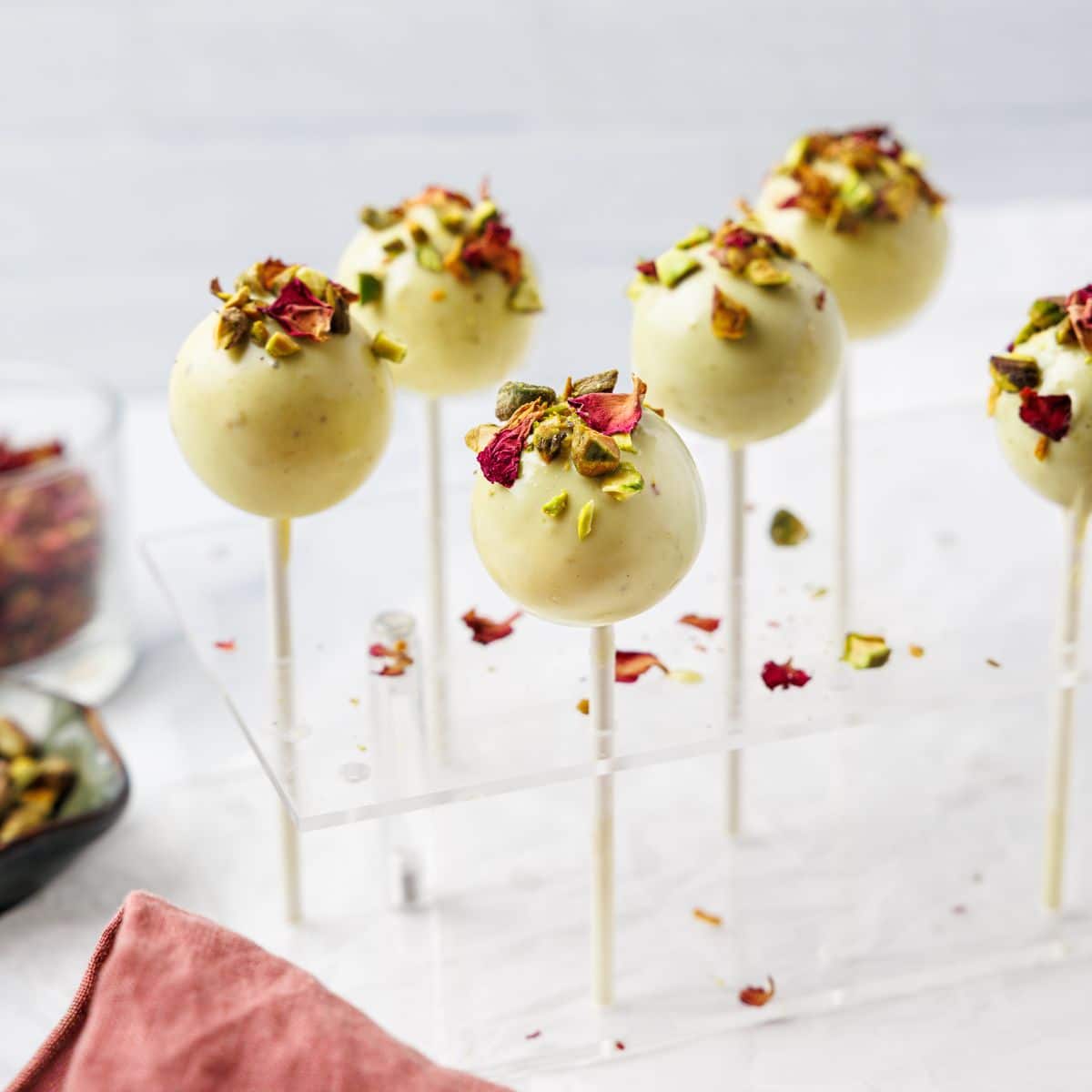 Pistachio cake pops coated in white chocolate topped with chopped pistachios and dried rose petals displayed in acrylic cake pop stand