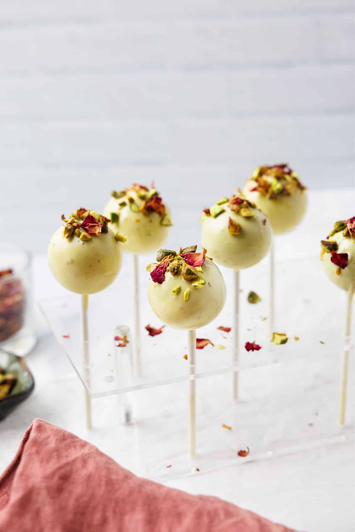 Pistachio cake pops on sticks displayed in acrylic stand, coated in white chocolate with crushed pistachios and rose petals