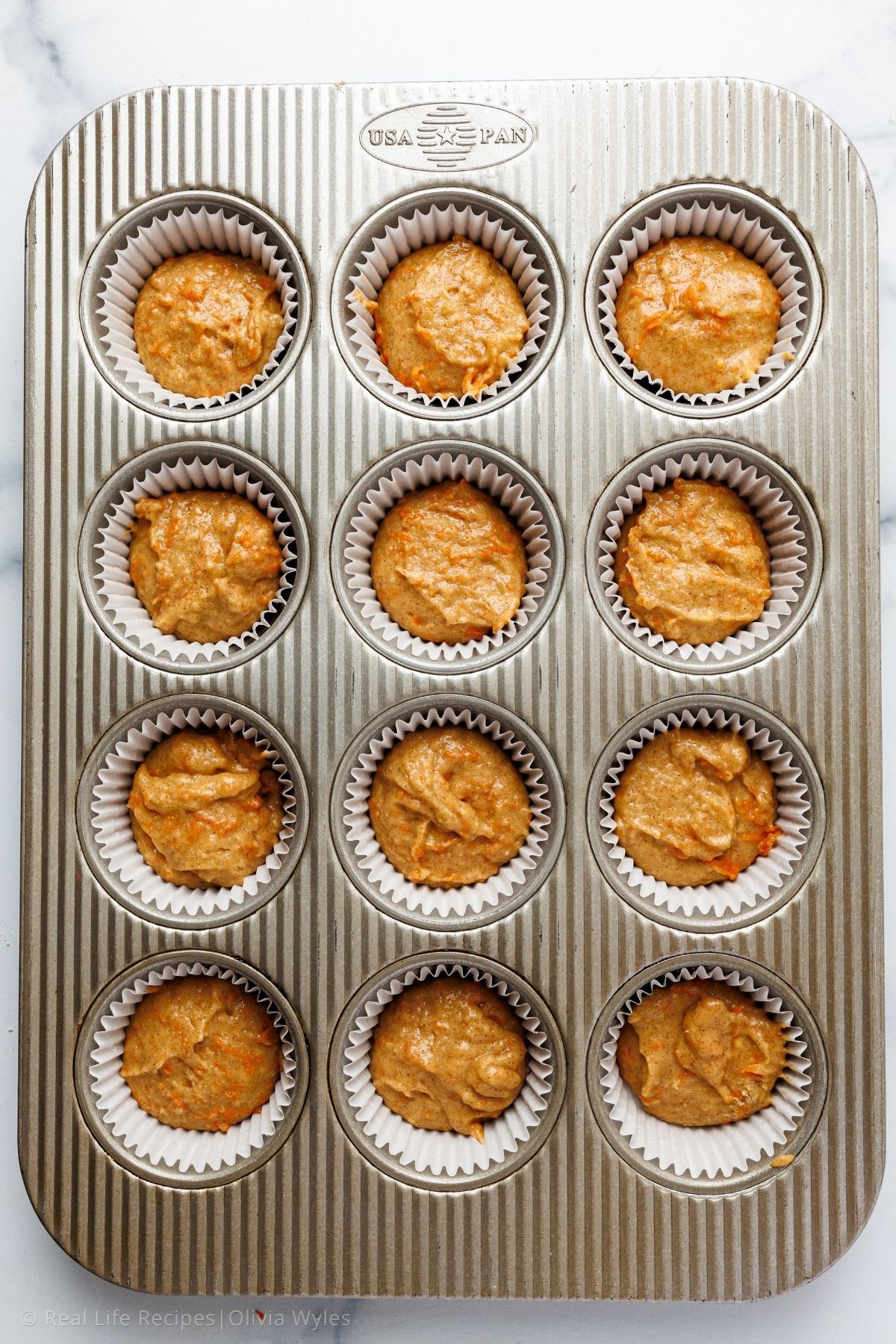 Twelve cupcake liners filled with carrot cake batter in a muffin tin, ready for the oven