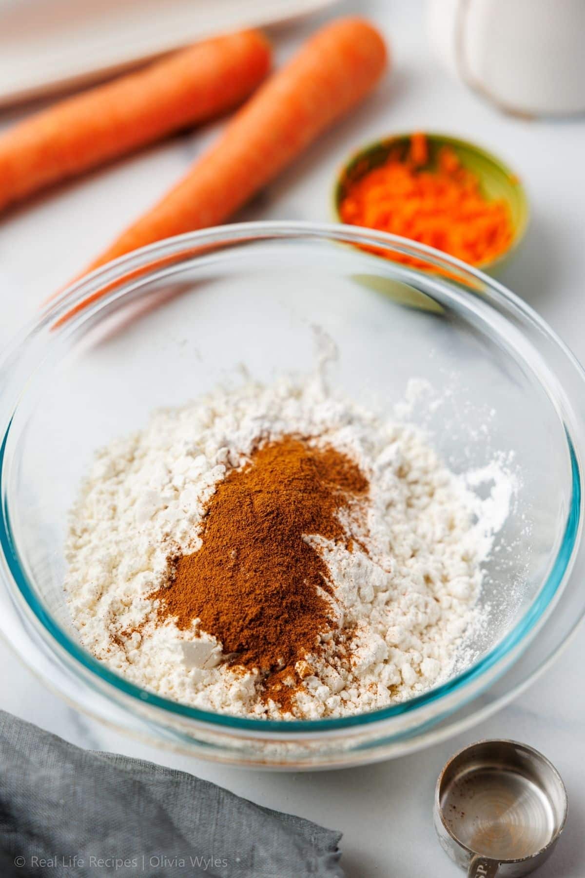 Almond flour, protein powder, baking powder, and warm spices measured into a glass bowl with fresh carrots nearby