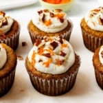 Six frosted carrot cake cupcakes topped with cream cheese frosting, chopped pecans, and finely grated carrots
