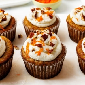 Six frosted carrot cake cupcakes topped with cream cheese frosting, chopped pecans, and finely grated carrots