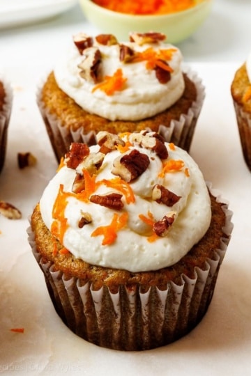 Six frosted carrot cake cupcakes topped with cream cheese frosting, chopped pecans, and finely grated carrots