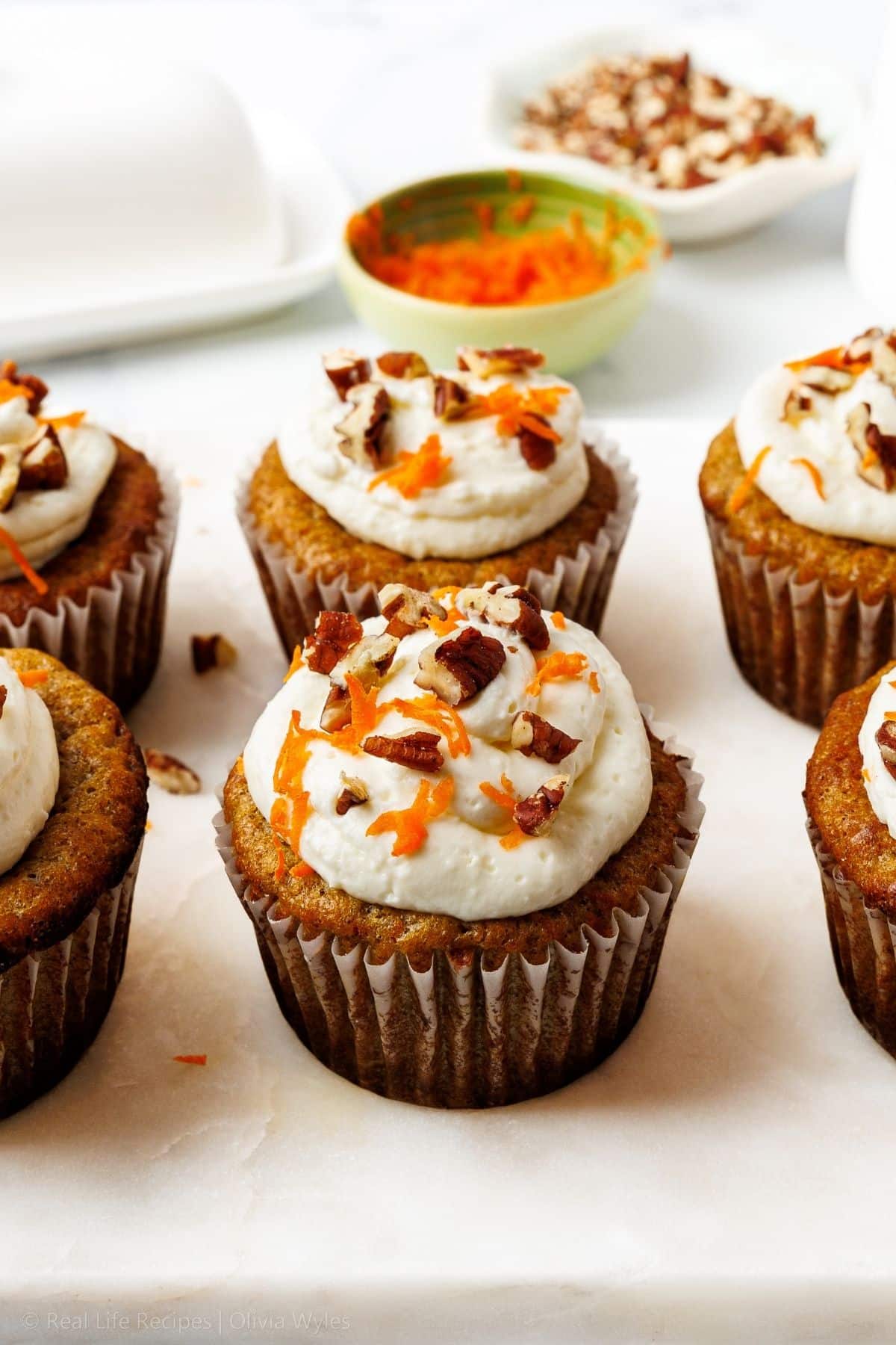 Keto carrot cake cupcakes with cream cheese frosting, chopped pecans, and finely grated carrot garnish on a marble surface