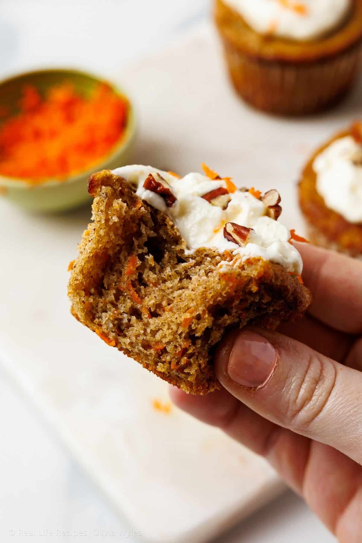 A hand holding a carrot cake cupcake with a bite taken out, showing the moist, tender crumb