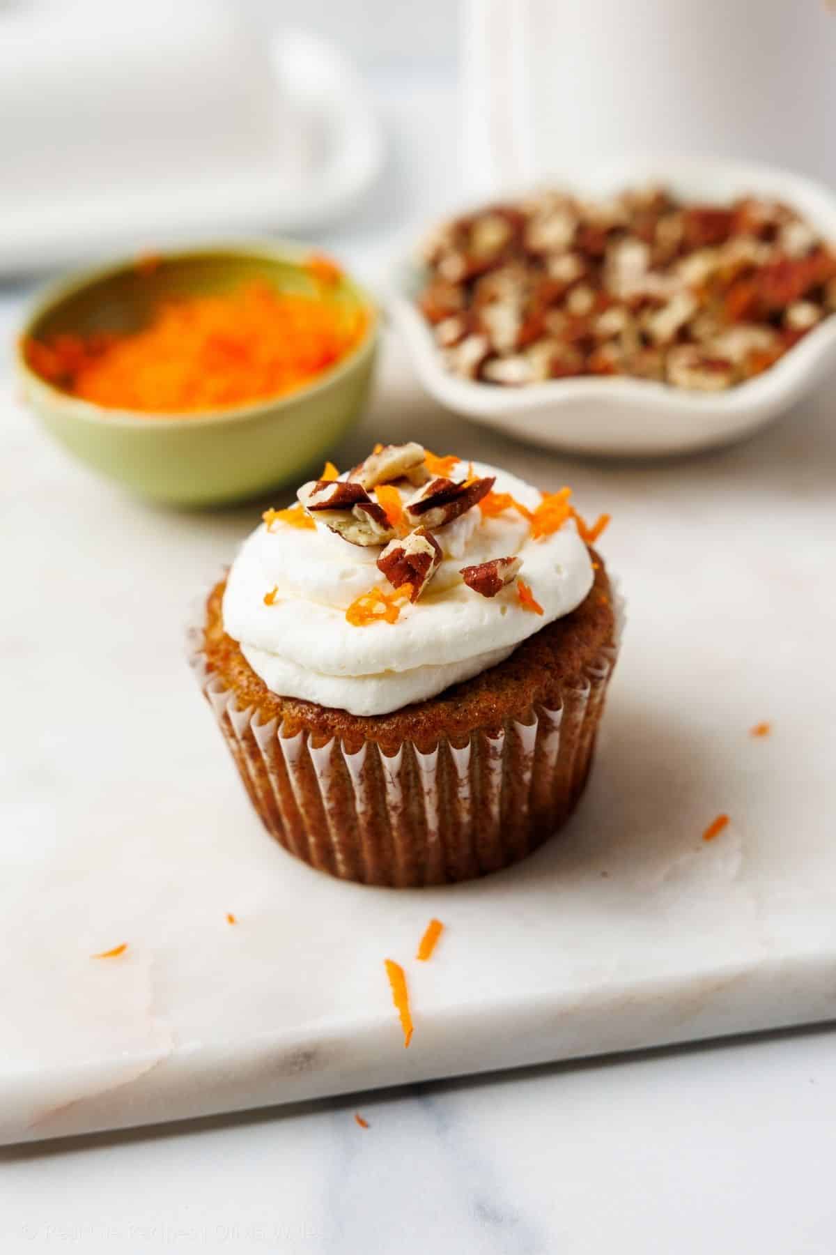 A single carrot cake cupcake with a swirl of cream cheese frosting, finely grated carrots, and chopped pecans on top