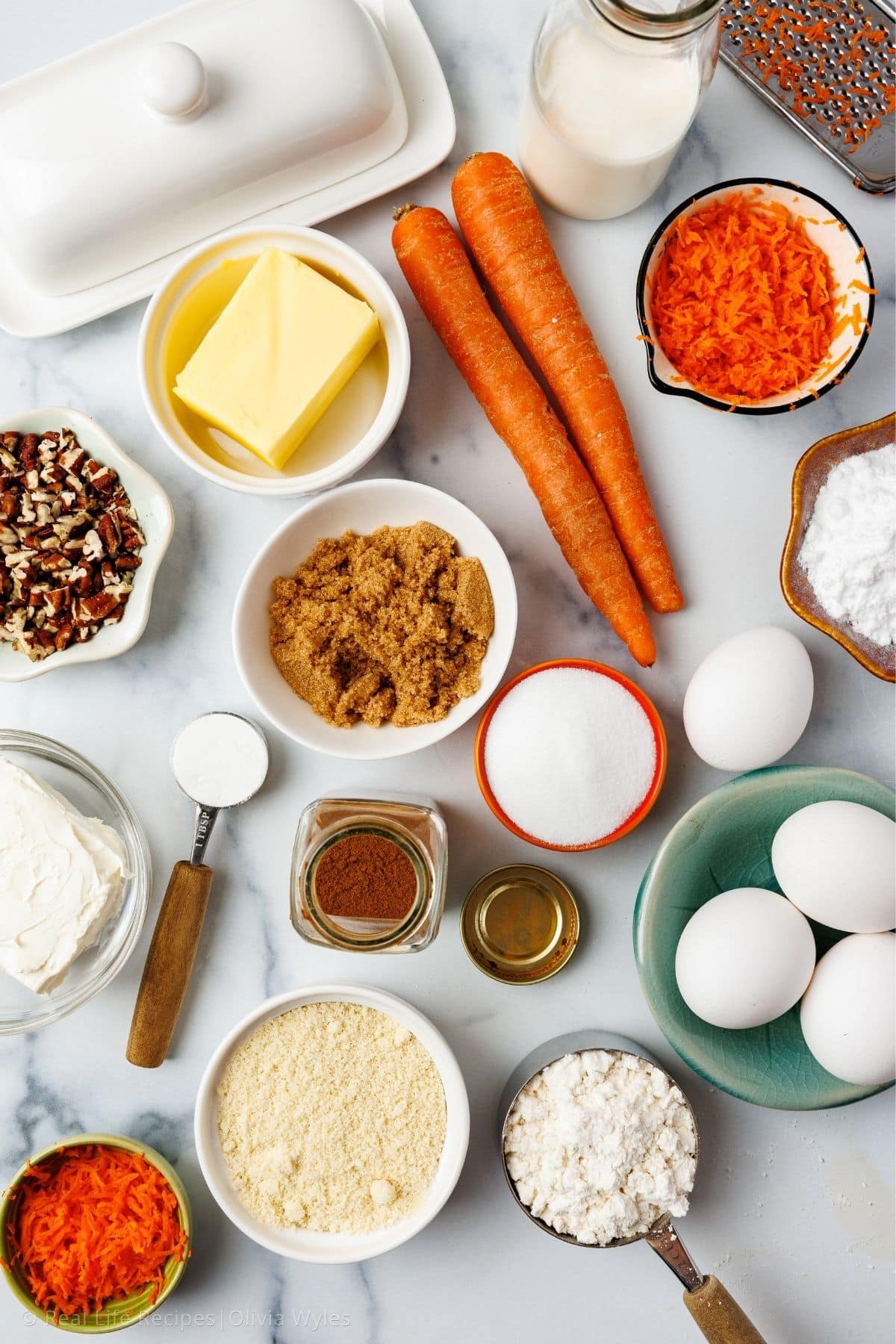 Overhead flat lay of all recipe ingredients including whole carrots, eggs, butter, almond flour, protein powder, cream cheese, and pecans