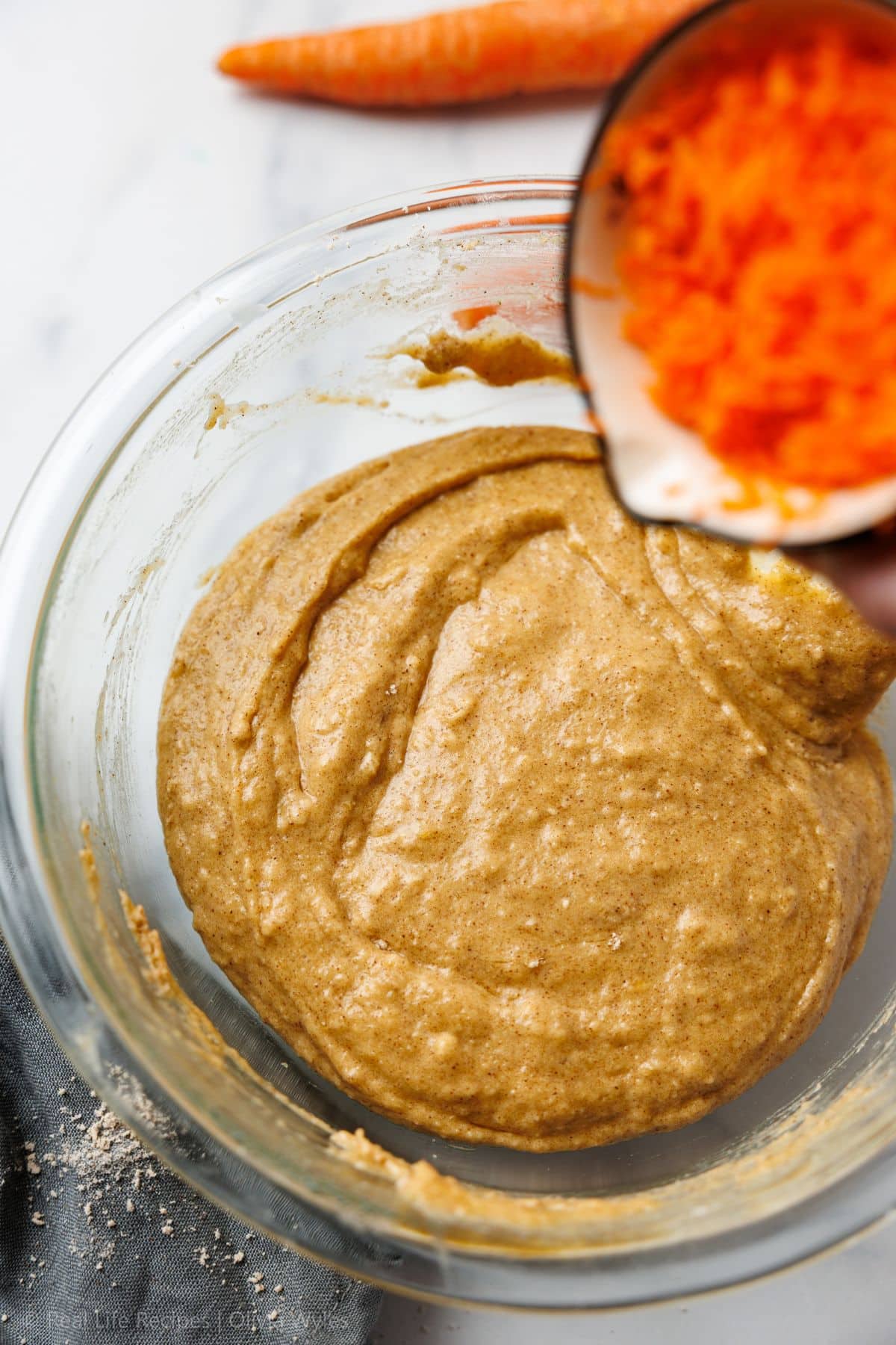 Smooth cupcake batter in a glass bowl with finely grated carrots being poured in from a measuring cup