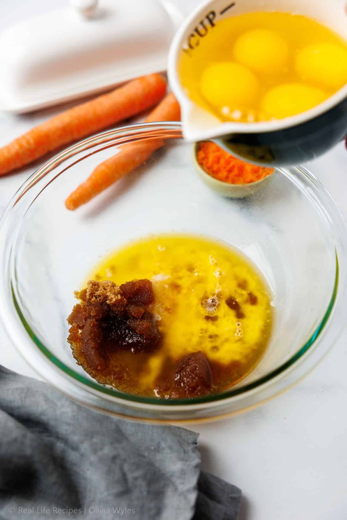Three eggs being poured over melted butter and brown sweetener in a glass mixing bowl
