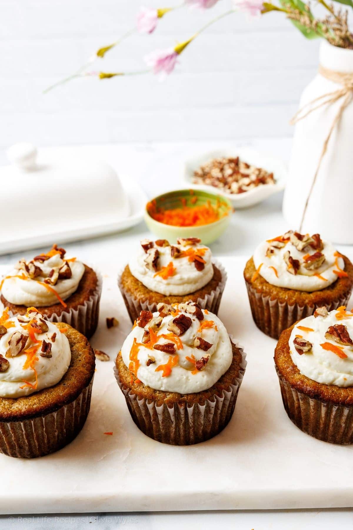 Six frosted carrot cake cupcakes on a marble board with small bowls of carrots and pecans and pink flowers in the background