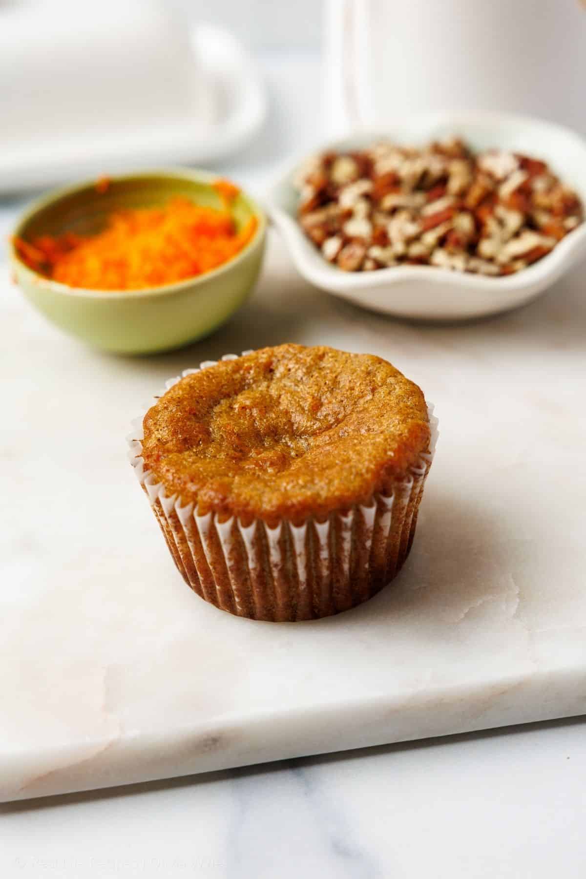 A single unfrosted carrot cake cupcake on a marble board with bowls of finely grated carrots and chopped pecans nearby