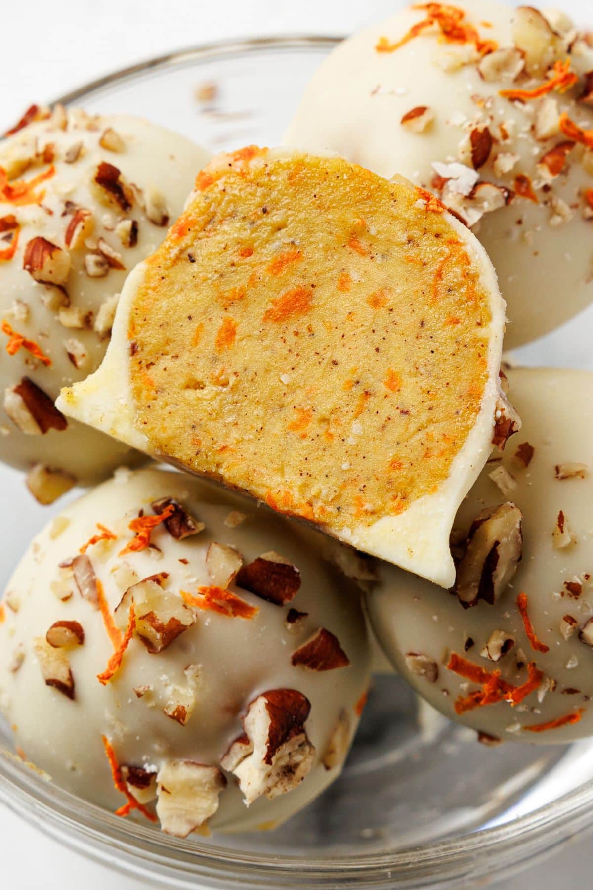 Keto carrot cake bites coated in white chocolate with pecans and shredded carrots, one bitten in half to show the spiced inside