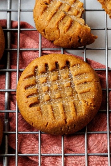 Single keto peanut butter cookie in focus on a wire rack over a rust napkin, with a deep criss-cross fork pattern.