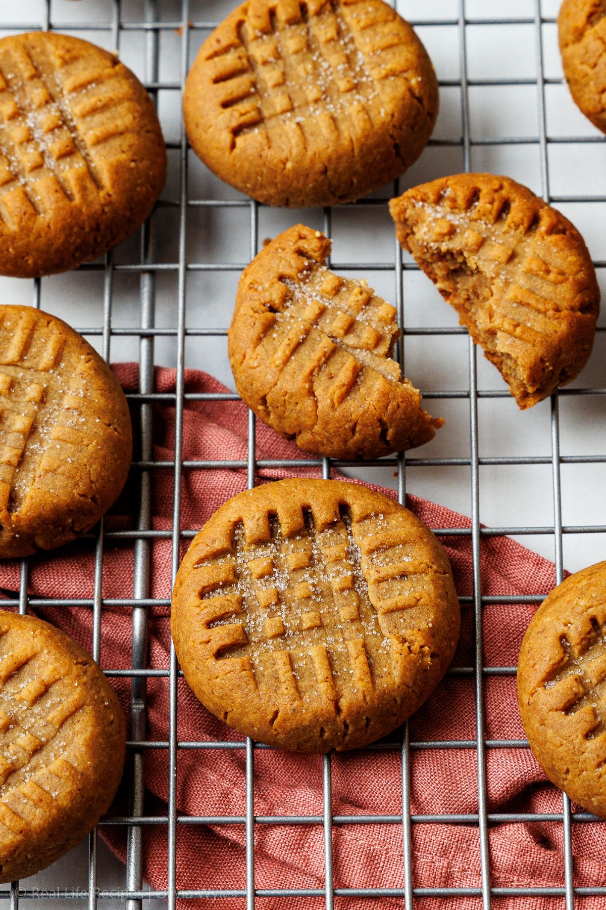 sugar-free peanut butter cookies scattered on a wire rack over a rust napkin, one broken in half showing soft interior.