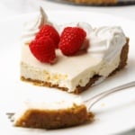 A slice of no-bake cheesecake on a white plate with whipped cream, raspberries, and a fork with a bite taken